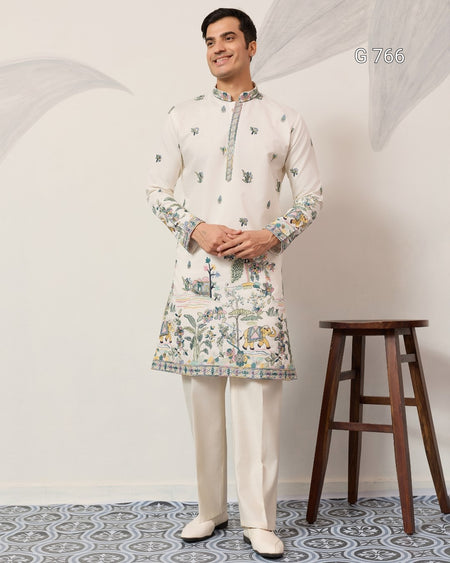 Man wearing a traditional kurta with floral patterns standing next to a wooden stool against a white wall.