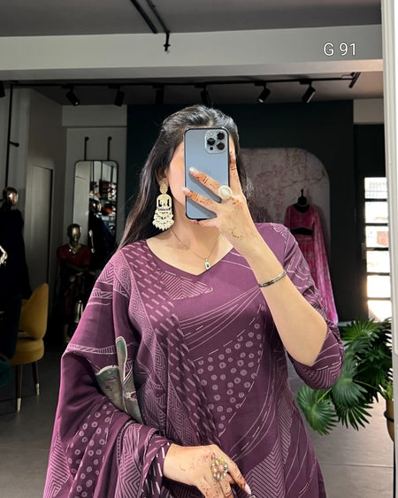 Woman taking a mirror selfie wearing a purple patterned outfit in an indoor setting.