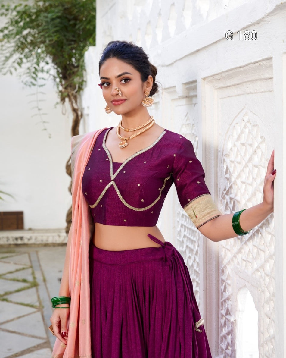 Woman in a purple traditional outfit with a pink dupatta standing against a white wall.