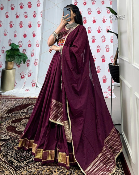 Person wearing a purple saree with gold border in a room with a decorative wall.