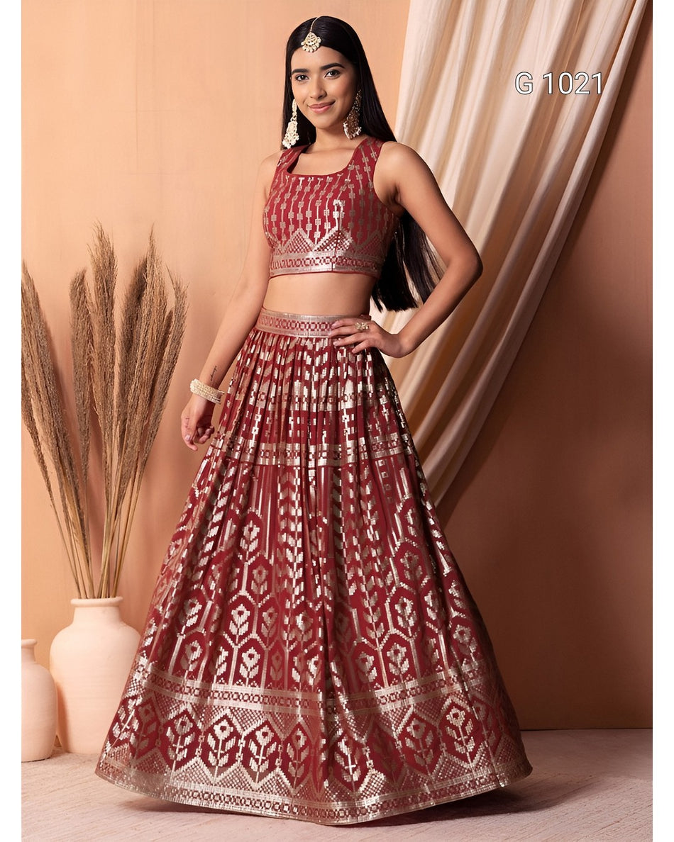 A woman models a traditional Indian lehenga choli set in rust red with intricate gold metallic patterns. The set includes a sleeveless crop top and a full flared skirt with geometric and floral motifs.