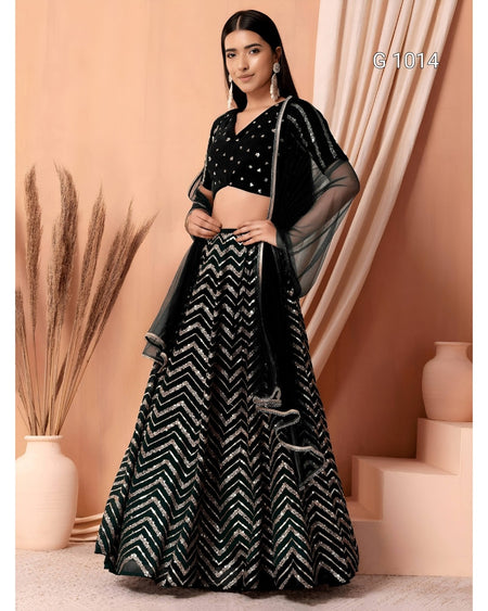 A woman models a black and silver embellished Indian ethnic wear set, featuring a flared skirt with a silver chevron pattern, a black embroidered crop top, and sheer black dupatta, set against a peach-colored backdrop with decorative vases.