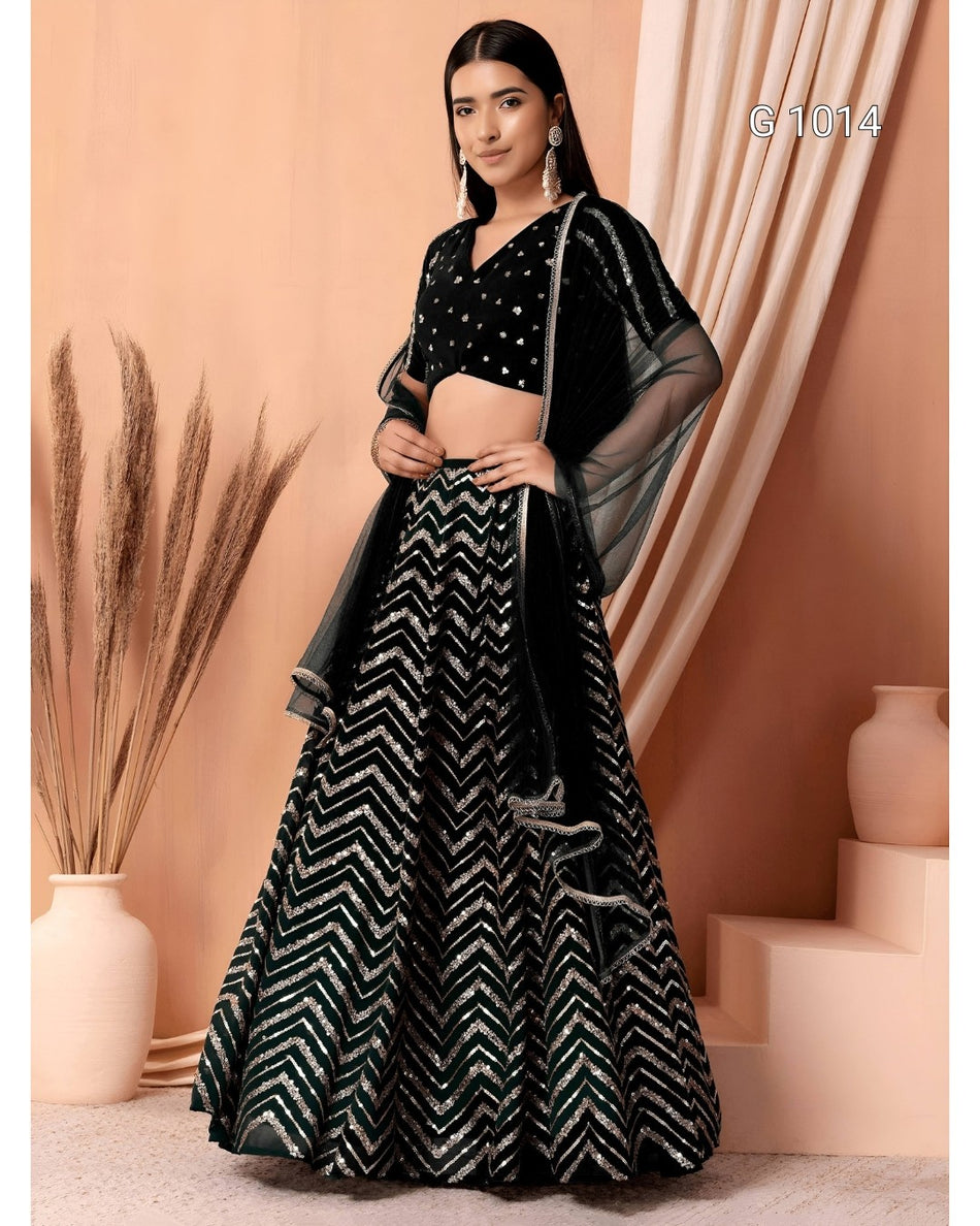 A woman models a black and silver embellished Indian ethnic wear set, featuring a flared skirt with a silver chevron pattern, a black embroidered crop top, and sheer black dupatta, set against a peach-colored backdrop with decorative vases.