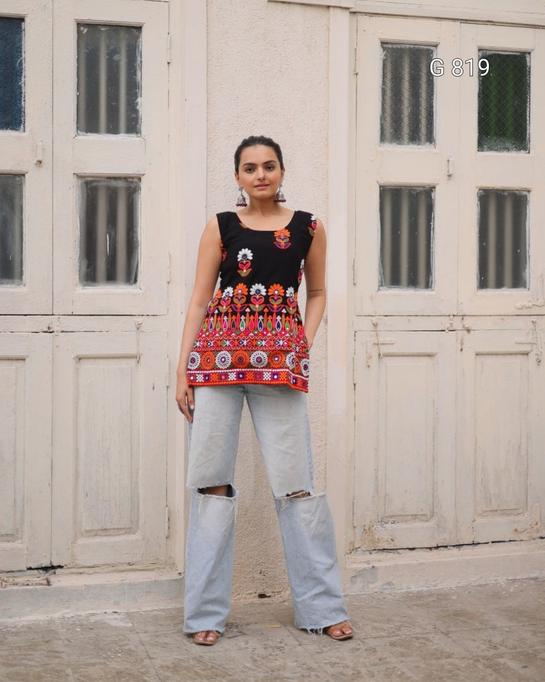 Woman wearing a colorful sleeveless top and light blue jeans standing in front of a white wooden door.