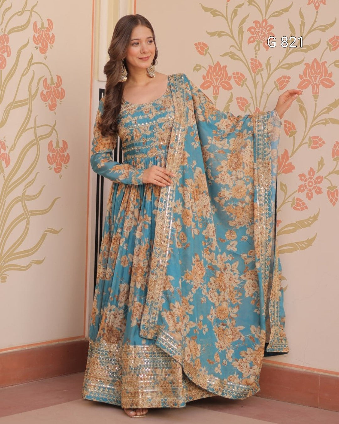 Woman wearing a blue and gold floral traditional outfit with a matching dupatta against a decorative wall.