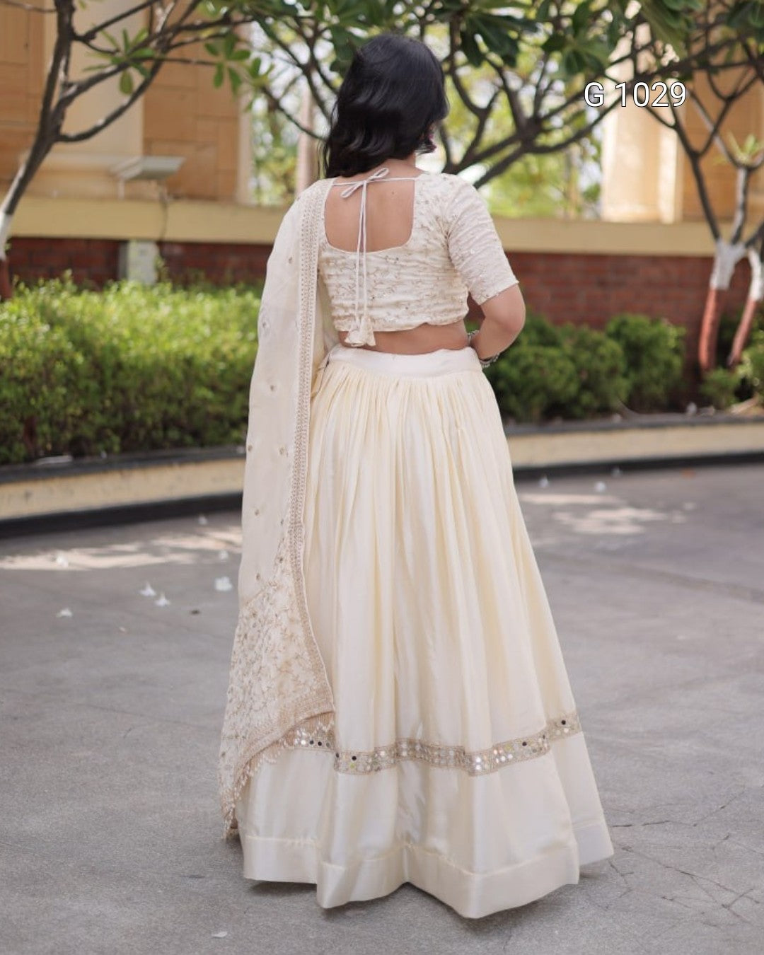 A woman shown from behind wearing a cream-colored traditional Indian outfit consisting of a lace blouse, flared skirt, and matching dupatta with mirror work, standing outdoors.