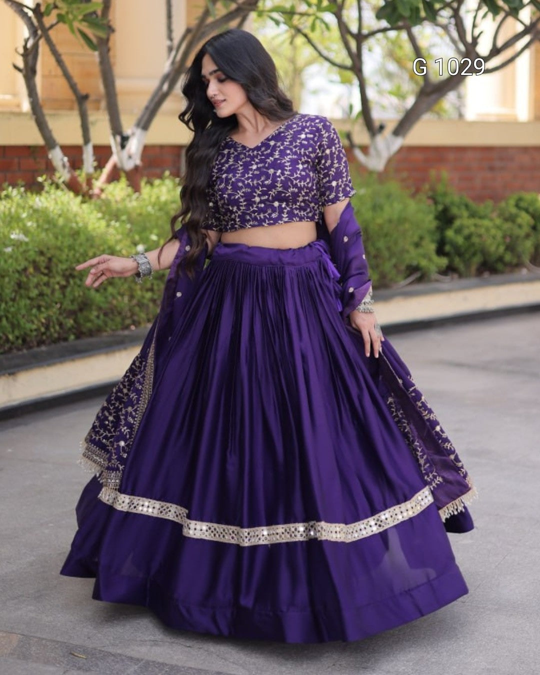 A woman modeling a purple Indian ethnic wear set consisting of a heavily embroidered crop top, a pleated flared skirt, and a matching sheer dupatta with silver mirror work borders, set against an outdoor garden background.