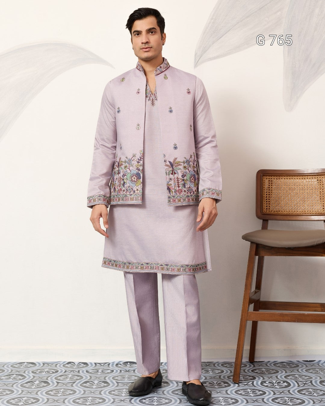 Man wearing a light purple embroidered traditional outfit standing in a room with a chair.