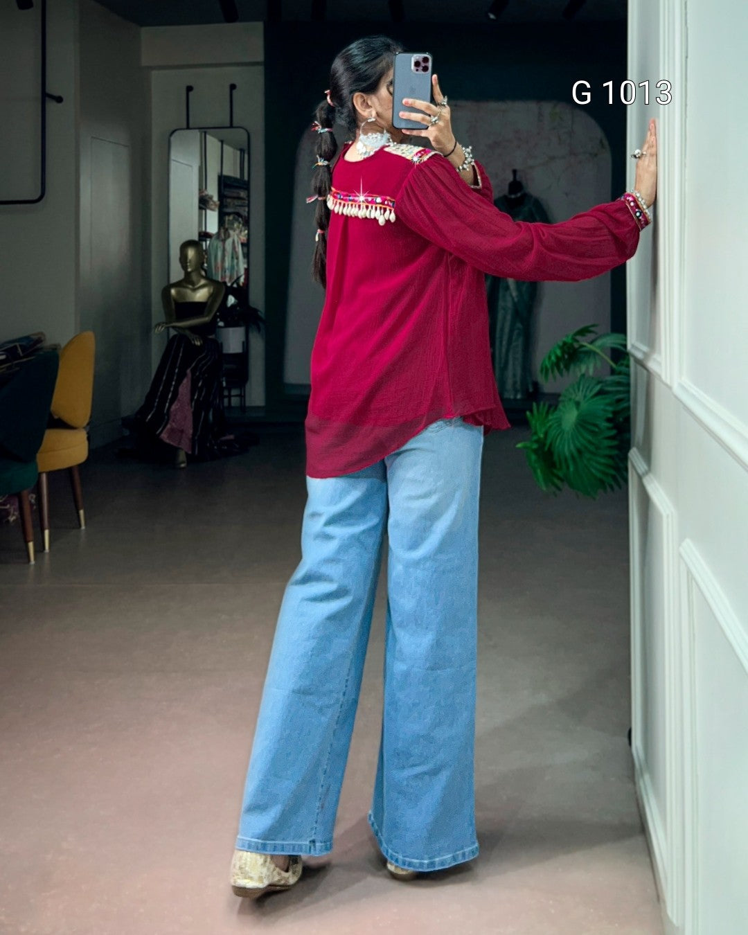 A woman takes a mirror selfie in a boutique, wearing a red long-sleeve blouse with gold embroidery and light blue wide-leg jeans. The boutique interior features a pinkish-brown floor, a yellow chair, and a white paneled wall.
