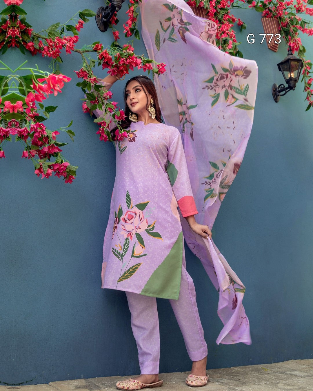 Woman in a lavender traditional outfit with floral embroidery against a blue wall with pink flowers.