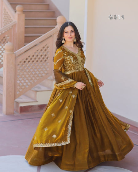 Woman in a mustard yellow traditional outfit with gold embroidery standing in a decorative interior setting.