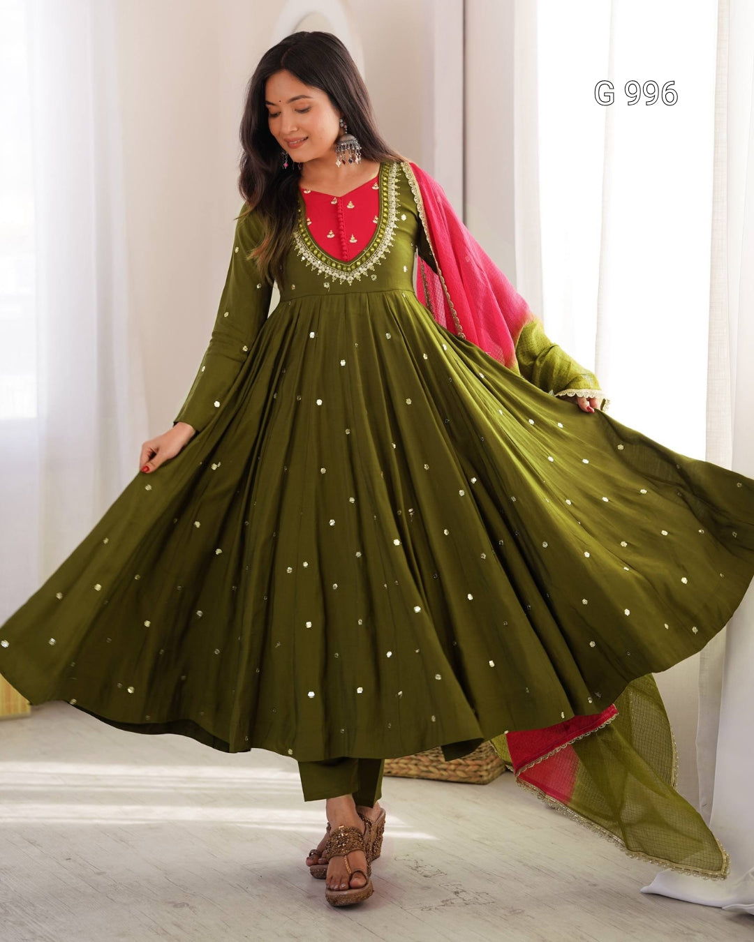A woman modeling a traditional olive green Anarkali gown with a red embroidered neckline, gold embellishments, and a bright pink dupatta, set against a soft white curtain background.