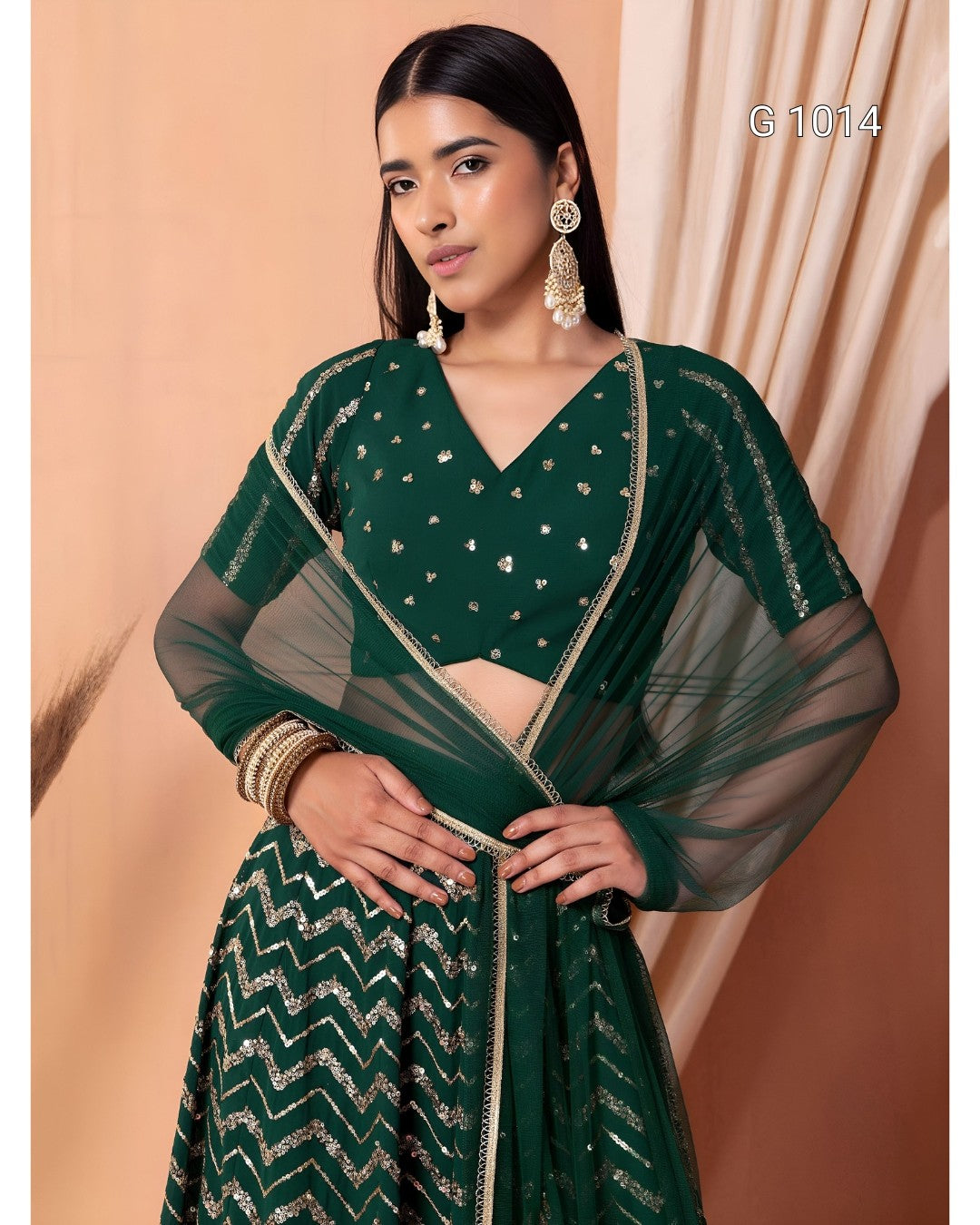 A woman modeling a dark green traditional outfit with a patterned skirt, sheer sleeves, and gold embellishments, posed against a peach-colored backdrop.