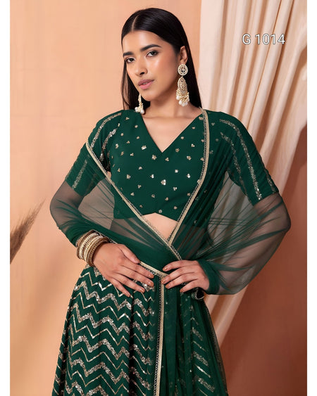 A woman modeling a dark green traditional outfit with a patterned skirt, sheer sleeves, and gold embellishments, posed against a peach-colored backdrop.