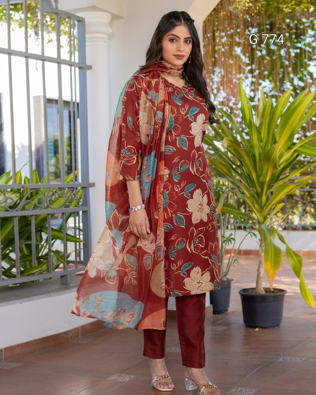 Woman wearing a red floral outfit standing outdoors with plants and a building in the background