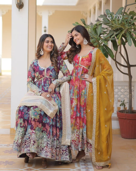 Two women in colorful traditional outfits standing indoors with a plant in the background.