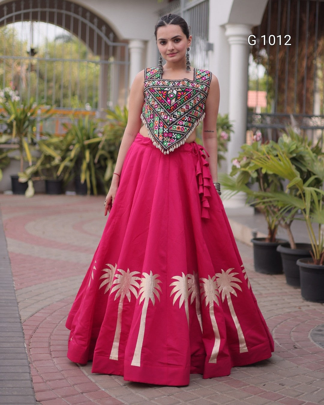 A woman modeling a bright pink traditional formal gown with a heavily embroidered sleeveless bodice and gold palm tree motifs near the hem, standing outdoors on a paved walkway.