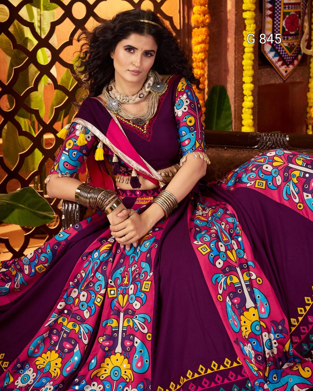 Woman in a colorful traditional outfit with intricate patterns, sitting against a decorative background.