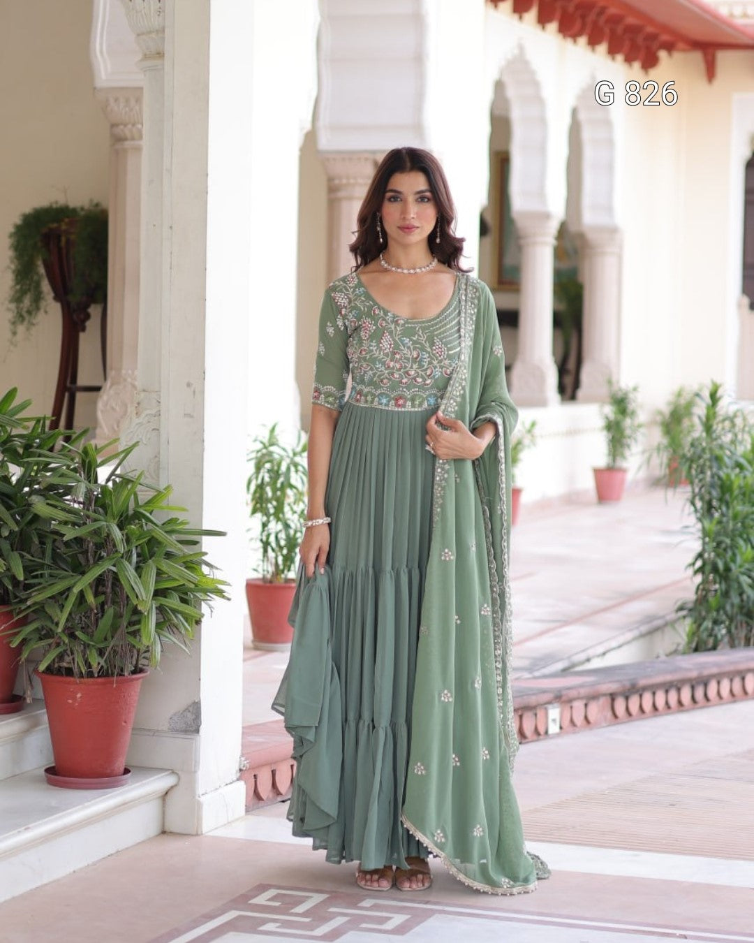 Woman in a green traditional outfit standing in a decorative outdoor setting with plants and architectural elements.