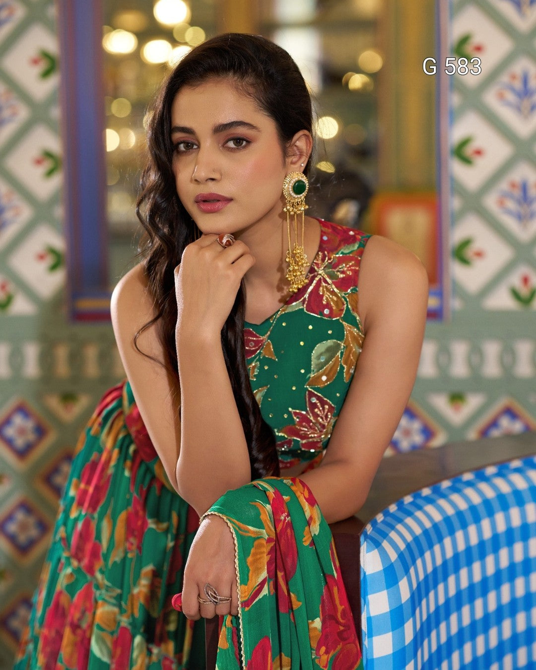 A woman wearing a sleeveless green saree with a colorful floral print and gold embroidery, paired with long gold earrings, posed in front of a patterned wall.