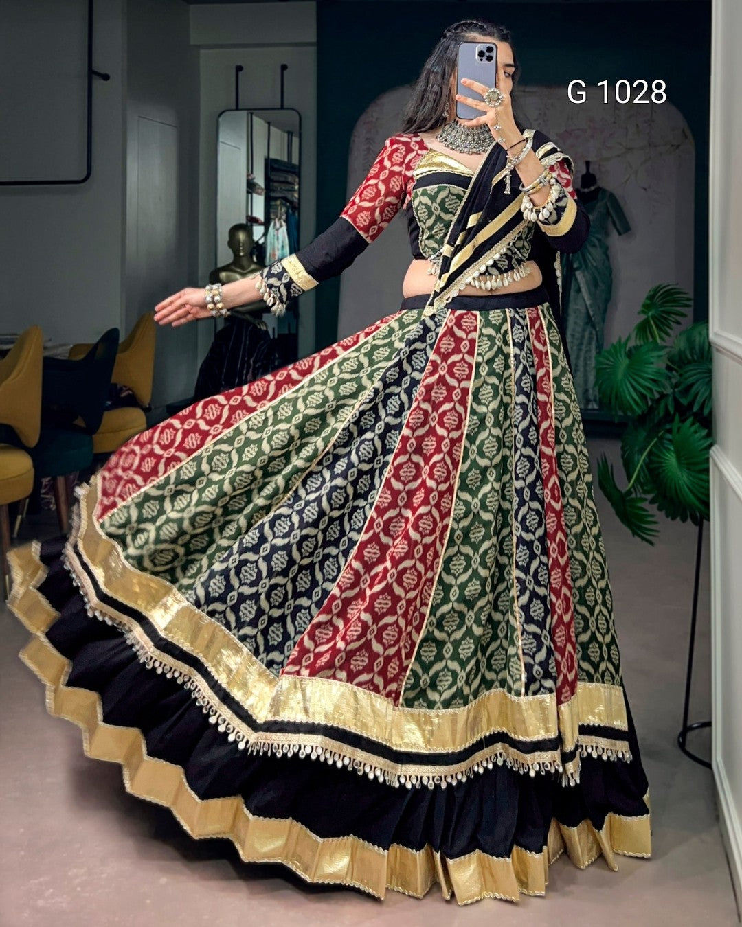 A woman takes a mirror selfie wearing a traditional Indian lehenga choli set. The skirt features vertical panels of red, black, and green with intricate gold and white floral patterns, and is finished with a wide gold border.