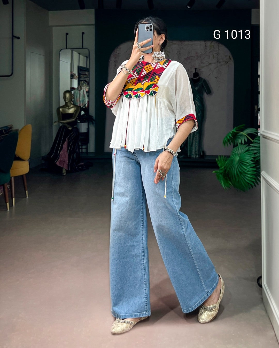 A woman takes a mirror selfie in a boutique wearing a white blouse with colorful geometric embroidery on the chest and sleeves, paired with wide-leg light blue jeans and metallic gold shoes. The boutique interior is visible in the background, and the text 'G 1013' is printed on the wall.