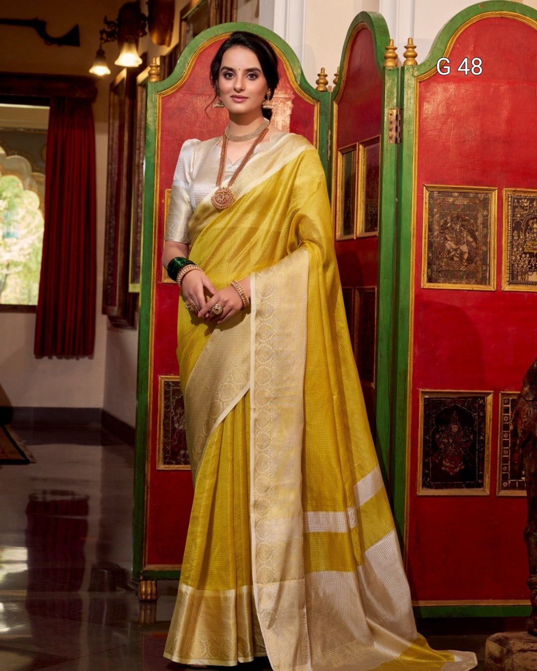 Woman in a yellow saree standing in a decorated room with red walls and gold accents.