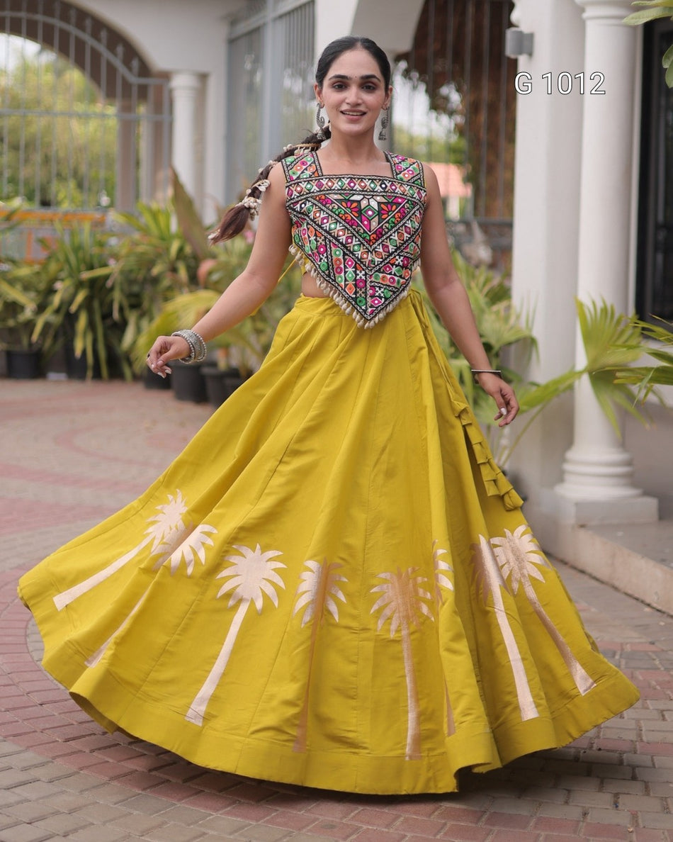 A woman wearing a bright yellow traditional Anarkali gown with a heavily embroidered multicolor bodice and white palm tree motifs near the hem, standing outdoors in front of a white columned building.