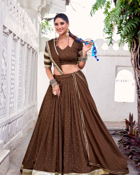 Woman in a brown traditional outfit with gold details standing in a decorative indoor setting.