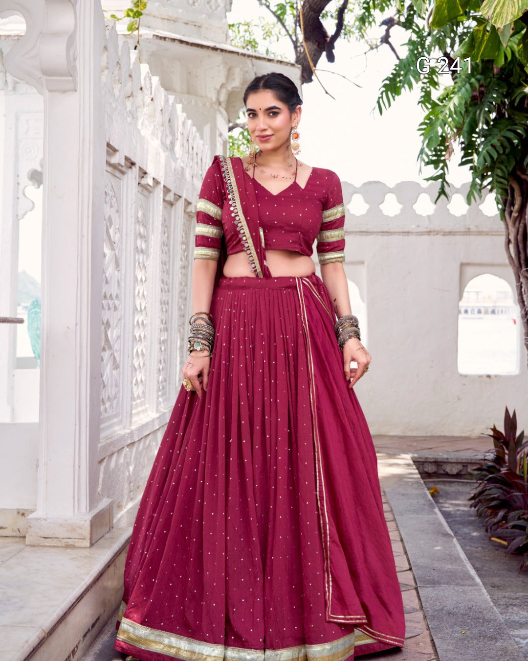 Woman in a maroon traditional outfit standing outdoors with white architecture and greenery.