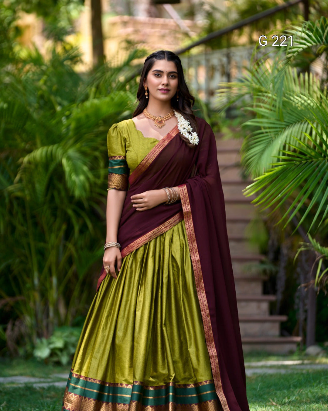 Woman in a green and maroon saree standing outdoors with greenery in the background