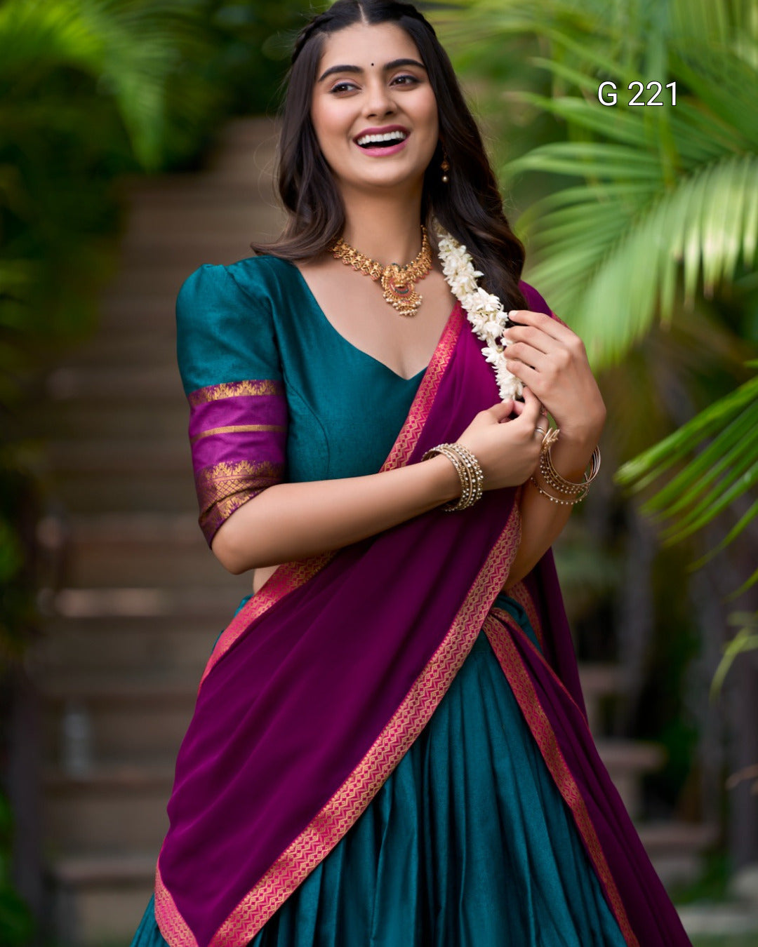 Woman in a traditional teal and purple saree with greenery in the background