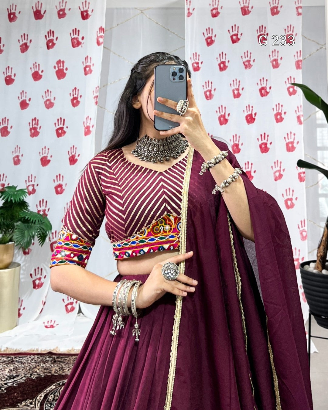Person wearing a maroon traditional outfit with colorful patterns, standing in front of a white curtain with red handprints.