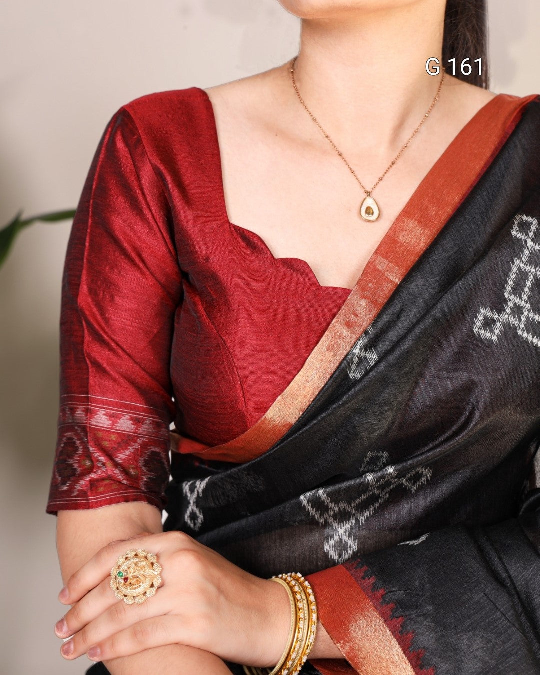 Person wearing a red blouse with a black saree, gold jewelry, and a blurred background.