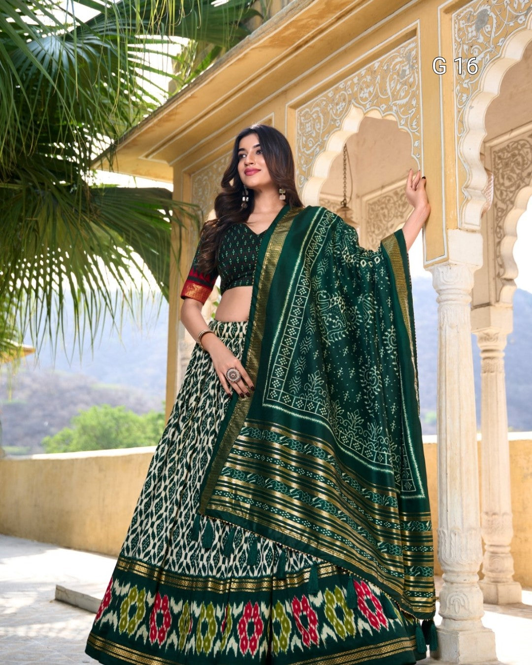 Woman in a green and gold traditional outfit holding a beige clutch, standing in front of an ornate building.
