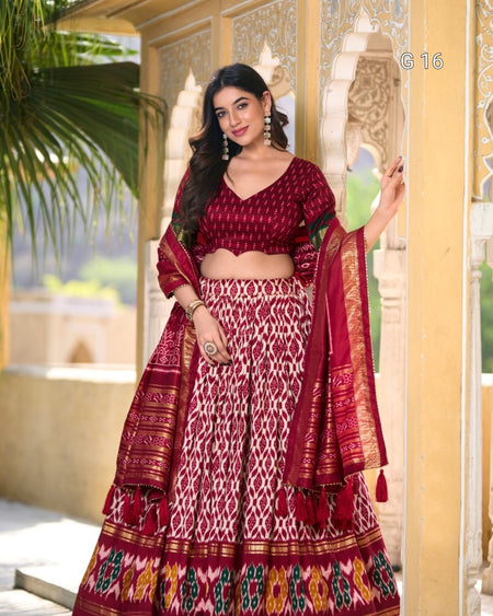 Woman in traditional maroon and white saree with intricate patterns, standing in a decorative setting.