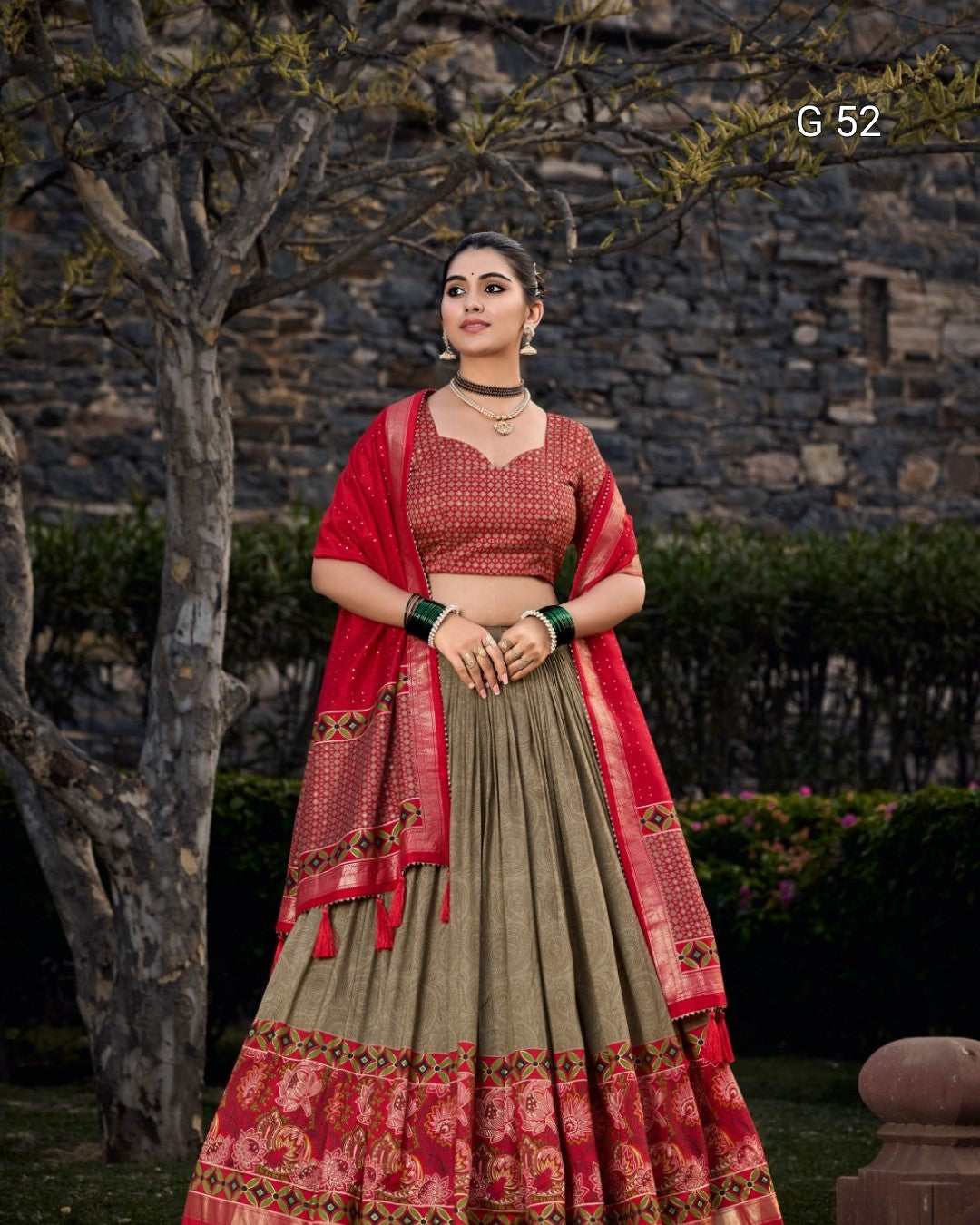 Woman wearing a traditional red and green outfit with a stone wall and trees in the background