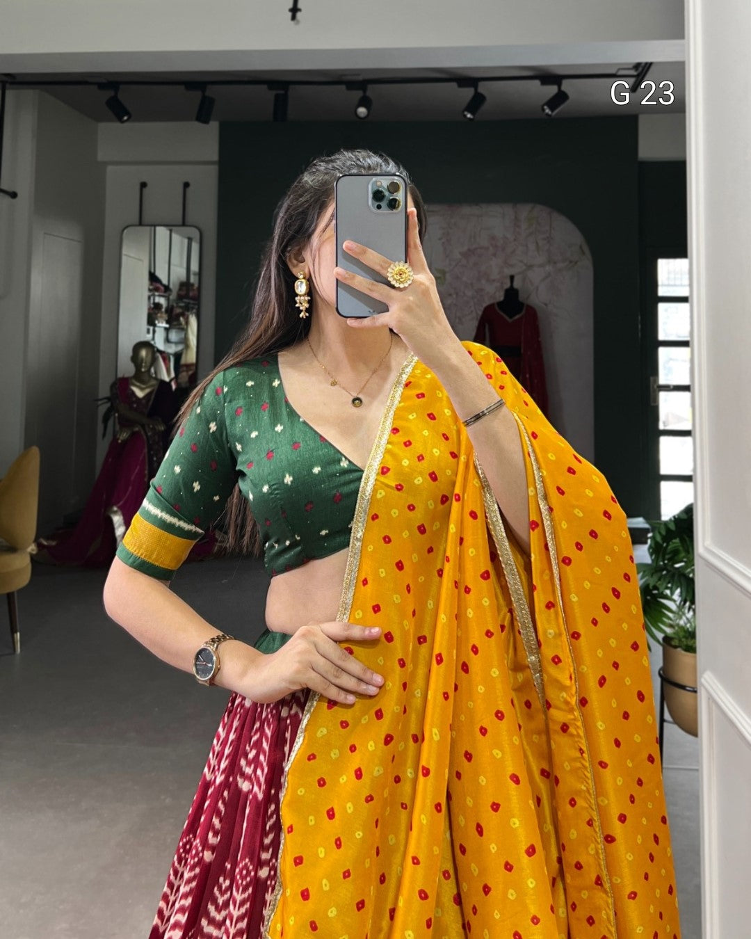 Person wearing a green blouse with gold trim and a yellow saree with red patterns, taking a mirror selfie.